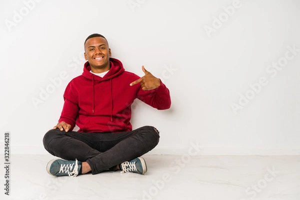 Fototapeta Young latin man sitting on the floor isolated person pointing by hand to a shirt copy space, proud and confident