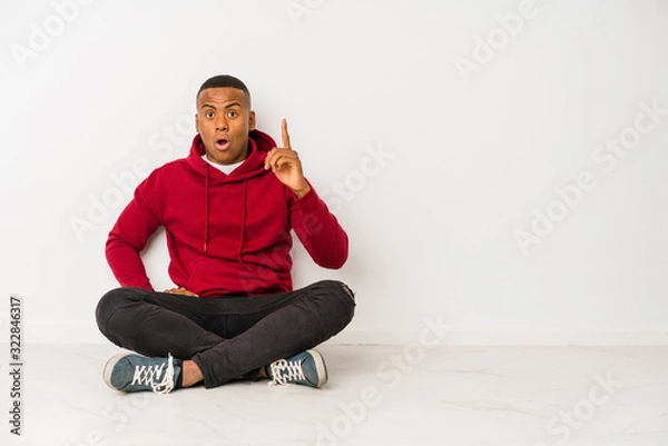 Fototapeta Young latin man sitting on the floor isolated having an idea, inspiration concept.