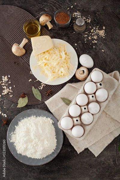 Fototapeta Ingredients for cooking, flour, eggs, grated cheese, mushrooms and spices, on a black background. Concept of making food, recipes.