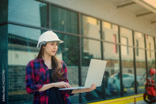Fototapeta A female engineer is analyzing the building structure plan on a portable computer and she is feeling the heat from the air in front of the construction project.