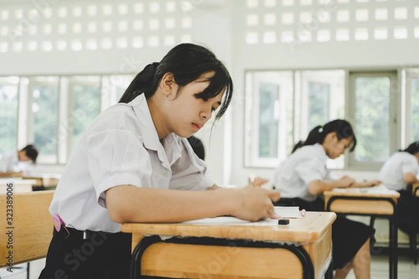 Fototapeta Teen young Asian students group concentrate one's mind writing test in exam on paper answer sheet and sitting on taking final examination ,in rows desk at classroom with Thai student uniform