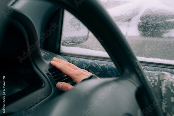 Fototapeta man's light-skinned hand is warming near a car heater in the black interior of the car against the background of a window in the snow. Warming up the car in bad weather in winter.