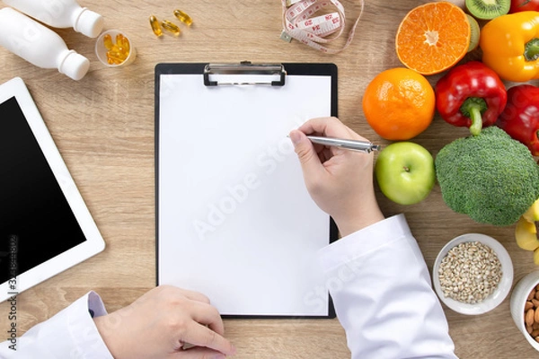 Fototapeta Top view of nutritionist doctor writing meal plan, medical records on clipboard with fresh fruits, vegetables, vitamins, measuring tape and tablet on wooden table. Healthy diet and nutrition concept.