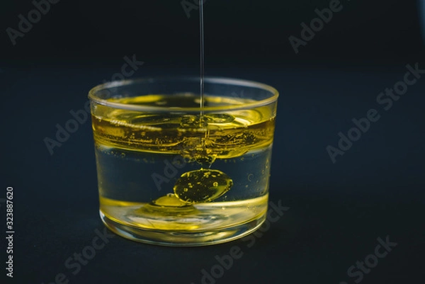 Fototapeta Olive oil falling on water in a low glass with drops of oil on the bottom. Isolated on a black background.Abstract concept.Copy space
