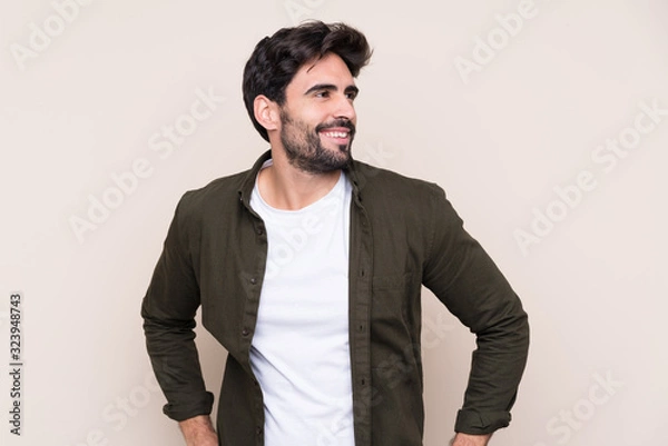 Fototapeta Young handsome man with beard over isolated background posing with arms at hip and smiling