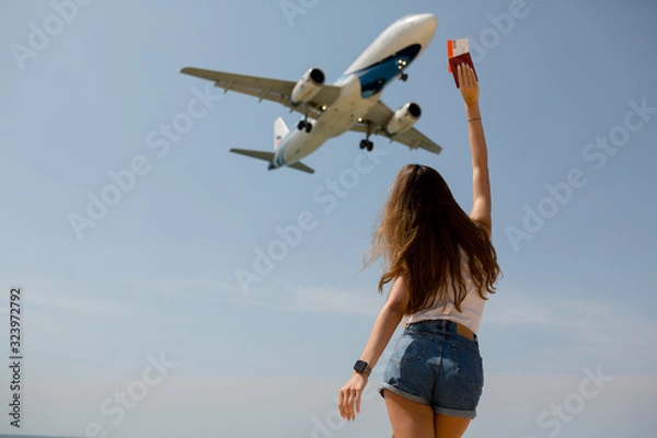 Obraz a slender girl in denim shorts and a white t shirt has her arms outstretched passport in hand near the airport and is watching the plane land in anticipation of a trip to Thailand