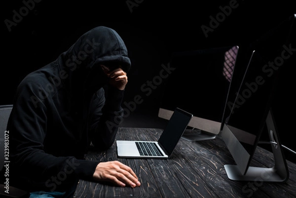 Fototapeta upset hacker sitting near laptop and computer monitors with blank screen isolated on black