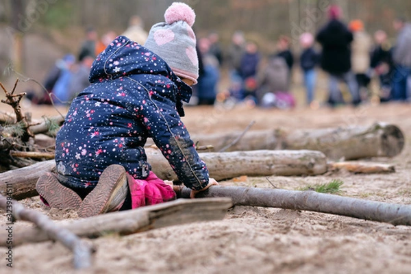Obraz Rear view of a 4 year old child girl in warm clothing kneeing on the ground  and building something with wooden branches in front of a group of people. Seen in Germany in February
