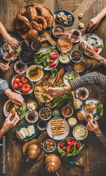Fototapeta Turkish breakfast. Flat-lay of peoples hands taking and eating Turkish pastries, vegetables, greens, cheeses, fried eggs, jams and tea in copper pot and tulip glasses over wooden background, top view