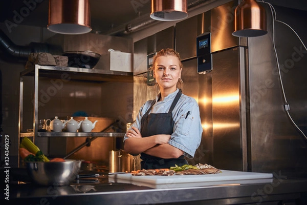 Obraz Confident and serious female chef standing with hands crossed in a dark kitchen next to cutting board with vegetables on it, wearing apron and denim shirt, looking in the camera, cooking show look