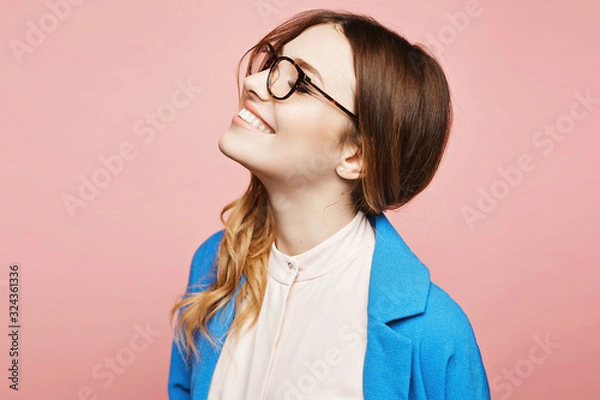 Obraz Happy young woman in stylish glasses and trendy blue coat, isolated at pink. Cheerful model girl in pink blouse and blue coat, and modish eyewear over pink background, isolated. Copy space. Fashion