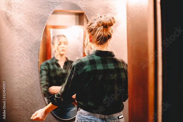 Fototapeta Young confident woman looking at herself in the mirror while standing in hallway at home getting ready before going out