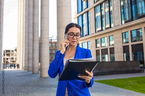 Obraz Confident woman skilled banker reading financial information from paper documents and talking via cell telephone while standing outdoors near contemporary enterprise building