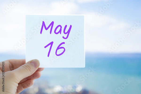Fototapeta May 16th. Hand holding sticker with text May 16 on the blurred background of the sea and sky. Copy space for text. Month in calendar concept
