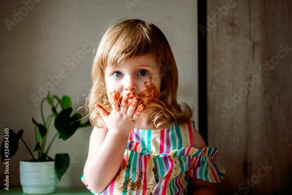 Obraz Cute curly haired baby girl eating chocolate,child smeared. dirty cheeks, nose and hands. Chocolate celebration on July 7th. harmful sweets for children. unhealthy food. indoor plants.
