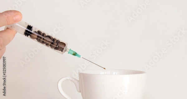 Obraz in female fingers a syringe with coffee beans over a coffee cup. At the end of the needle is a drop of coffee. Concept - Coffee Addiction