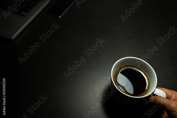 Fototapeta hand holding a white cup of hot black coffee against dark background with laptop and mobile phone