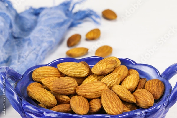 Obraz Almond nuts lie in an oval-shaped blue glass bowl and on a white surface, close-up