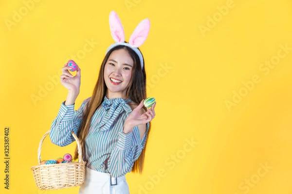 Fototapeta Asian young woman wearing bunny ears showing colorful easter eggs, hold basket filled eggs, toothy smiling happy face. Holidays and people concept isolated on yellow color background, copy space.