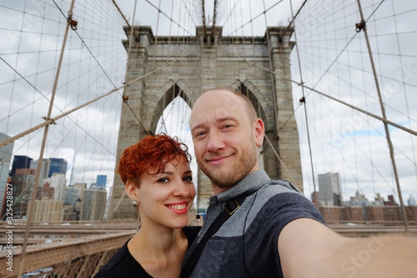 Fototapeta Happy selfie tourist woman and man taking self-portrait picture with smart phone on Brooklyn Bridge, New York City, Manhattan, USA. Young beautiful family couple travel, photographing NY landmarks.