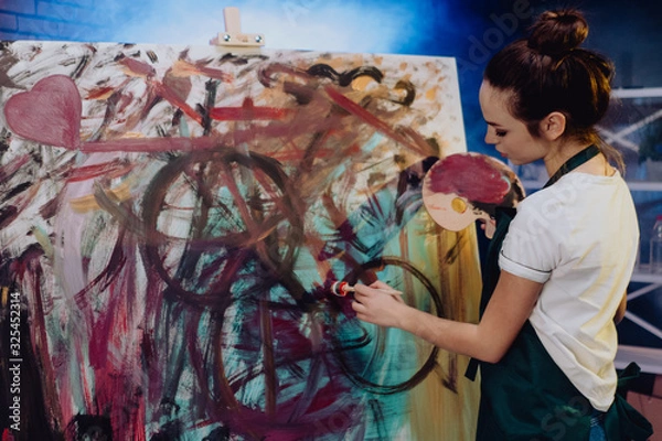 Fototapeta A young female artist is creating an abstract painting. She is working in the loft space with dimmed lights. She is using oil paint and a brush.