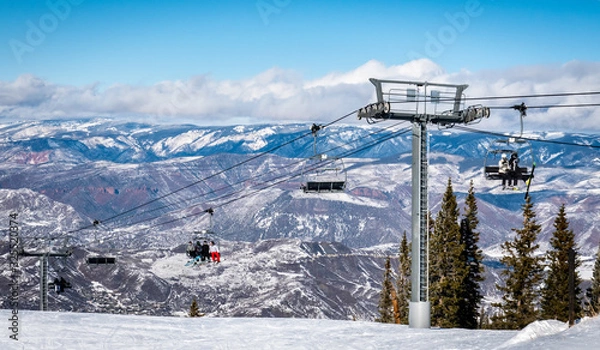 Fototapeta Skiers and snowboarders ascend the Elk Camp chairlift at the Aspen Snowmass ski resort, in the Rocky Mountains of Colorado on a partly cloudy winter day.