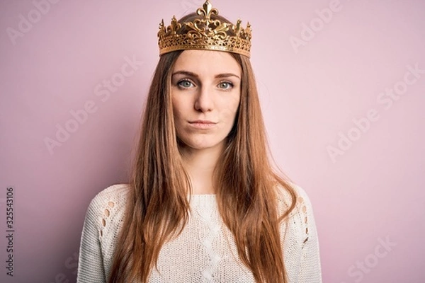 Obraz Young beautiful redhead woman wearing queen crown over isolated pink background with serious expression on face. Simple and natural looking at the camera.