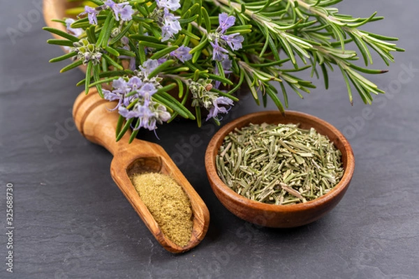 Fototapeta Purple blooming and fresh rosemary twigs in a bowl of olive wood, a small bowl with whole dried rosemary and a spice shovel with ground rosemary on a black slate background