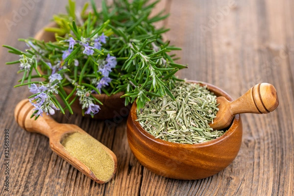 Fototapeta A wooden bowl with blooming and fresh rosemary twigs, a wooden bowl with whole dried rosmary and a spice shovel with ground rosemary on a rustic wooden background