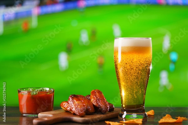 Fototapeta Spicy barbecue chicken wings, tomato salsa, nachos and beer on dark wooden bar table. Football on background, high resolution