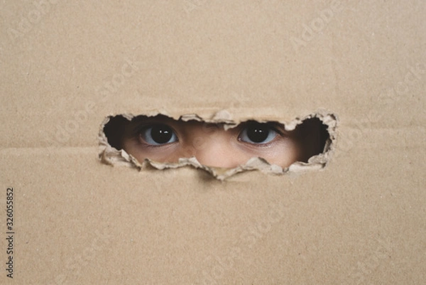 Fototapeta A child eye looking through a hole on a cardboard box. Concept of spy and stalker