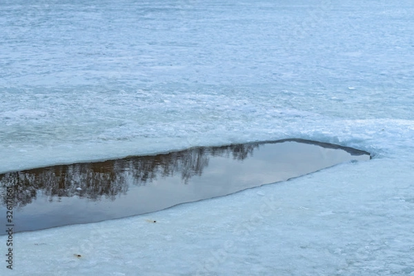 Fototapeta An ice hole in a frozen pond begins to melt. Trees and sky are reflected in the water. Spring is coming. Russia