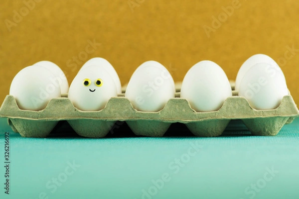 Fototapeta Happy smiling easter egg among simple white eggs in a paper tray on turquoise and mustard yellow bacground. Funny easter scene.
