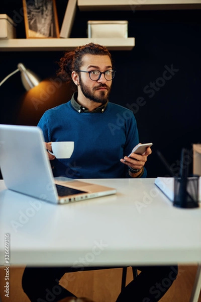Fototapeta Handsome bearded caucasian hipster sitting in his modern home office, using smart phone for checking messages on social media and holding cup of coffee.