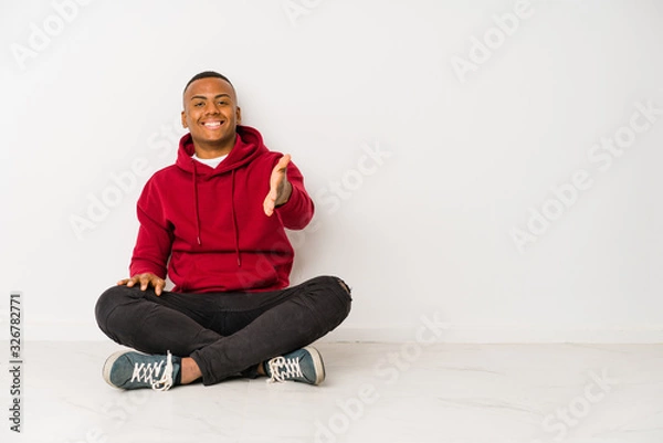 Fototapeta Young latin man sitting on the floor isolated stretching hand at camera in greeting gesture.