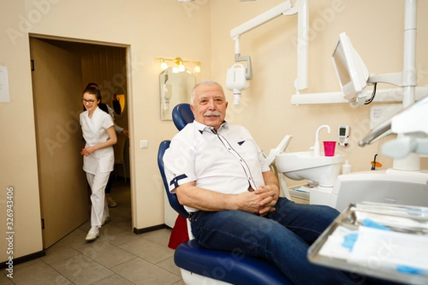 Fototapeta Portrait of happy senior man age 75 with mustache sitting in dentist’s chair, looking at camera while young dentist woman comes in the room to examine. Concept of dentistry, medicine and health care