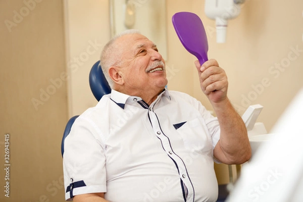 Fototapeta Closeup portrait of happy satisfied senior man looking at mirror and enjoying his beautiful toothy smile after treatment. Dental care for old people. Concept of dentistry, medicine and health care.