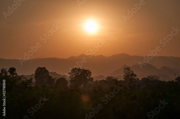 Fototapeta silhouettes of trees and layered mountains in an orange light with a setting sun in the background