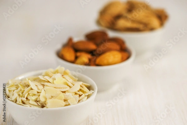 Obraz Coconut flakes and almonds in three white bowls on a white surface