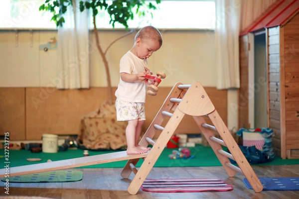 Fototapeta Child standing on a Pikler triangle with a red toy car in a playroom