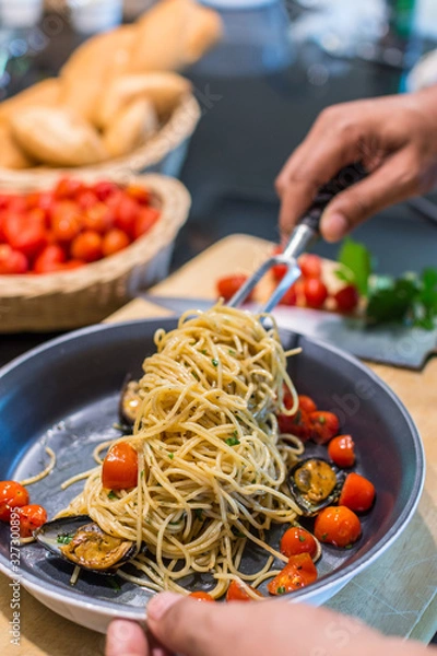 Fototapeta closeup of a chef forking up spaghetti Vongole with roma tomatoes and mussels in a pan - with a knife and cutboard with tomatoes and bread in the background