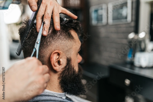 Fototapeta Good looking bearded man visiting hairstylist in barber shop.