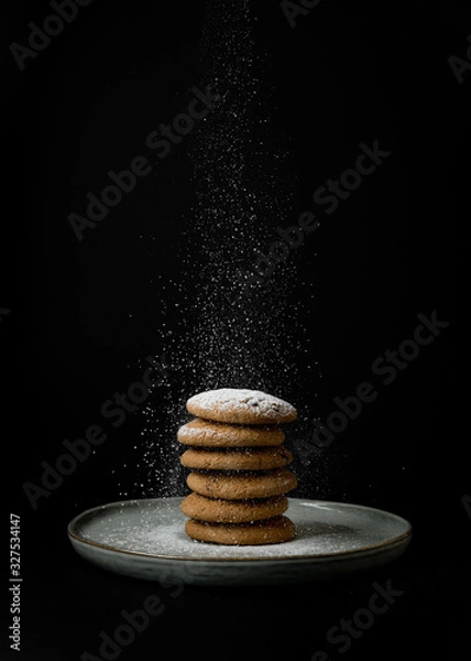 Fototapeta Sprinkling sugar powder stack of oatmeal cookies on a clay plate on black background. Isolate