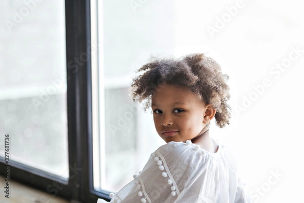 Fototapeta Close-up portrait of cute little african american kid girl in a white dress. She's posing sitting by the window.  Studio shoot. .