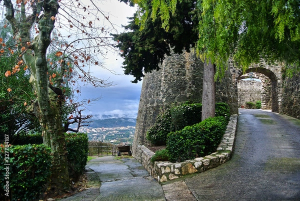 Obraz Saint-Paul-de-Vence / Provence - Alps - Cote d'Azur, France - Road on a hill with a stone fortified wall of the city, green trees and bushes, in the background a white-blue sky in the summer afternoon