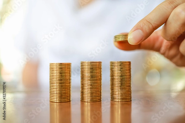 Fototapeta Business, Money, Finance, Saving and Secure Concept. Close up of man hand holding and put four coins to stack of coins on wooden table.