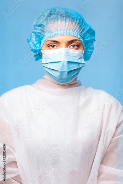 Fototapeta female doctor in medical uniform portrait, in a cap and a protective mask on her face. Blue background. Surgeon before a surgery.