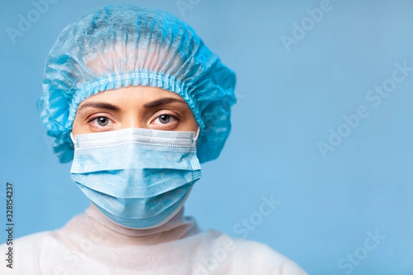 Obraz Portrait of a doctor, a young woman in a protective medical mask on her face and a cap on her head. looking seriously into the frame. on a blue background. surgeon. ambulance paramedic. copy space