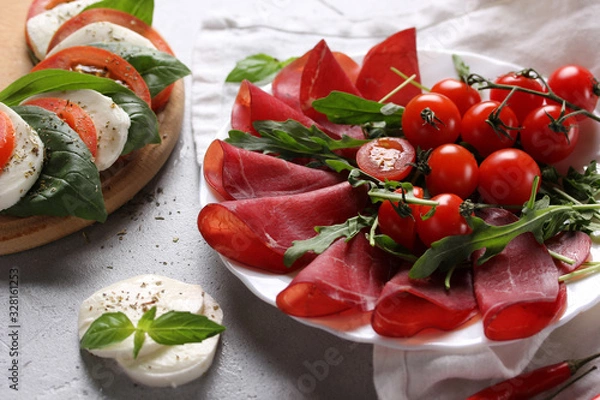 Fototapeta The concept of Italian cuisine. Italian dried ham from beef. Meat snack. Bresaola with arugula and tomatoes, mozzarella, basil and chili pepper. Caprese. Background image, copy space