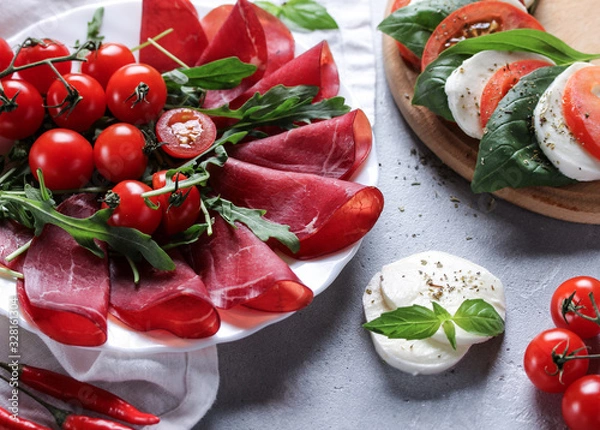 Fototapeta The concept of Italian cuisine. Italian dried ham from beef. Meat snack. Bresaola with arugula and tomatoes, mozzarella, basil and chili pepper. Caprese. Background image, copy space